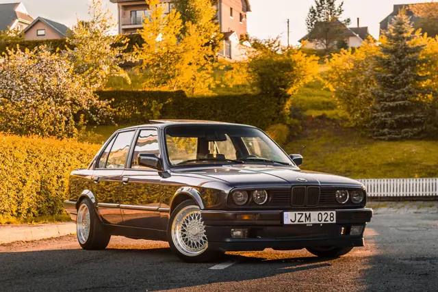 Side profile of a black BMW E30 sedan parked on a street with autumn trees behind